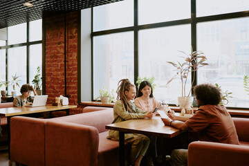 Group of four people working together in cozy cafe, with two women and two men sharing ideas using laptops and notebooks, large windows allowing natural light to flow in