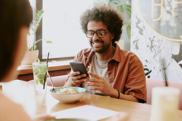 Smiling young man sitting at restaurant table interacting with smartphone while enjoying meal and beverage with friend sharing pleasant moment