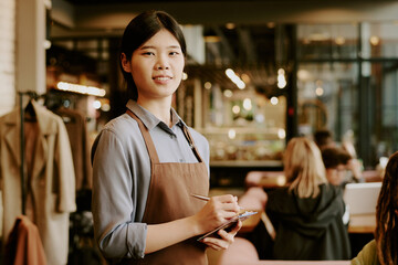 Smiling Asian waitress in apron holding notepad in busy cafe, engaging with customers. Background shows bustling cafe interior with patrons seated at tables and bar area