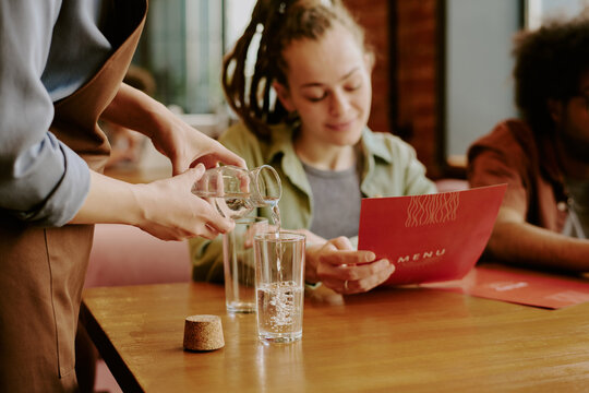 Server pouring water into glass for guest reading menu in restaurant with wooden tables and cozy ambiance. Interaction between server and guest creates warm, inviting experience