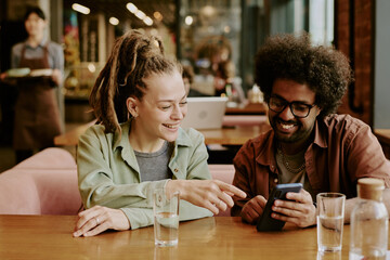 Two people laughing while looking at smartphone screen in casual cafe setting surrounded by tables, chairs, and indoor lights