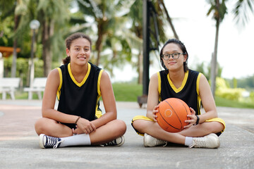 Two young girls wearing sports uniforms sit on ground with one holding a basketball. Background features lush green park with trees and benches