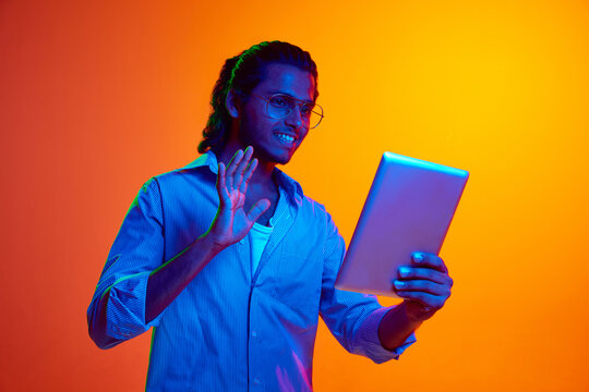 Smiling young middle eastern man waving hello calling his friends in distance in neon light against gradient orange background. Concept of human emotions, diversity, youth, modernity. Ad