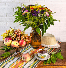 Still life with apples and grapes in a basket, a cup of tea, a bouquet of autumn flowers in a vase, a sun hat on a wooden background