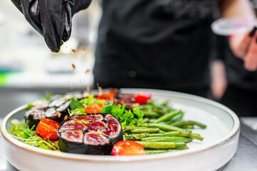 Close-up of a chef’s hands garnishing a gourmet dish with herbs. The plate features seared tuna, green beans, and cherry tomatoes, showcasing culinary artistry and attention to detail.