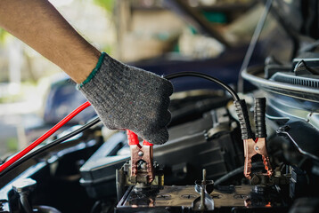 Auto mechanic’s hand holds a battery jumper cable to jump start a car with a dead battery.  Car...
