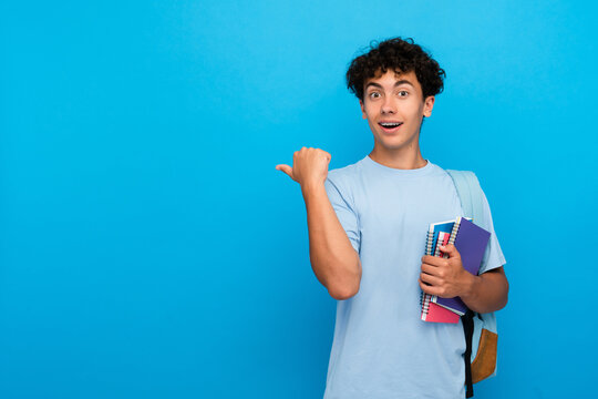 Amazed boy guy high school male pupil student teenager with bag holding books copybooks pointing finger empty copy space for advertisement isolated on blue background. Education back to school concept