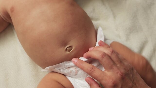 Unrecognizable mother massages newborn baby's tummy, gently touching child's navel, infant kid with naked body wearing only in diaper lying on white sheet