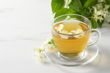 Aromatic jasmine tea in cup, flowers and green leaves on white marble table, space for text