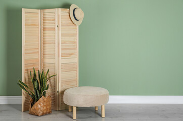 Wooden folding screen, pouffe, bag, houseplant and hat near green wall indoors, space for text