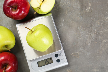 Kitchen scale with apple among ones on grey textured table, flat lay. Space for text