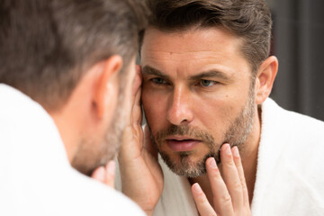 a man in a bathrobe in the bathroom looking at his wrinkles and pimples on his face. A man is looking at his reflection in a mirror. A man is looking at his reflection in a mirror. 