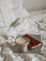 Aesthetic cozy morning in bed with striped linen - cappuccino cup, laptop, notebook