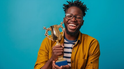 Happy male African American student boy raising a golden trophy posing in front of blue background with copy space, concept of winning, top prize, award winning prize.