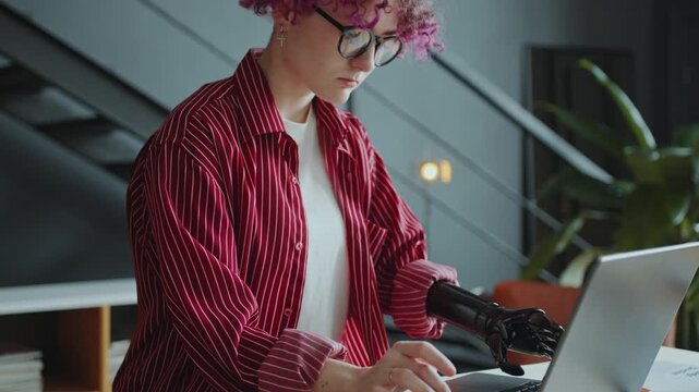 Curly haired girl with prosthetic arm using laptop and taking notes on paper at desk during workday in the office. Tilt-up shot