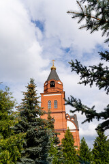 A charming red brick church featuring a prominent clock tower is beautifully surrounded by tall pine trees, creating a picturesque scene that enhances the aesthetic of the natural landscape