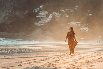 Beautiful brunette girl walking barefoot at the beach wearing a dress by sunset time