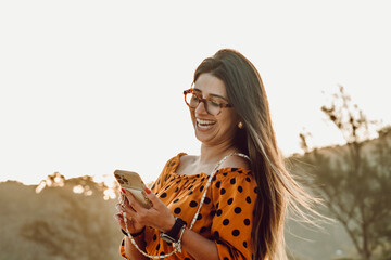 Beautiful brunette girl close-up shot smiling using her cell phone at the beach by sunset