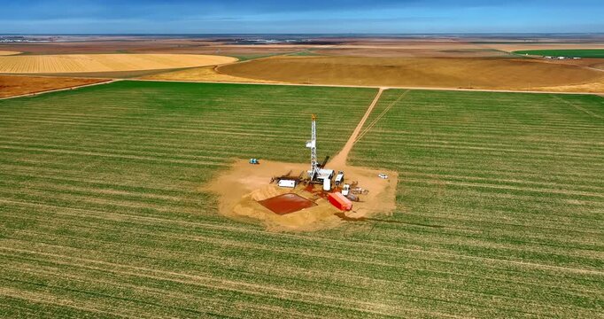 Big rectangular green field with drilling site in the middle. Drone footage around the oil production rig.