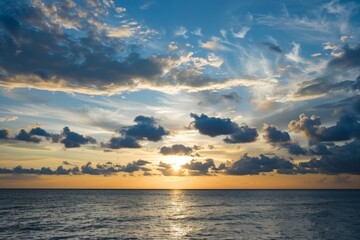 Obraz premium The sky is blue with clouds and the sun is setting. The ocean is calm and the water is a deep blue color