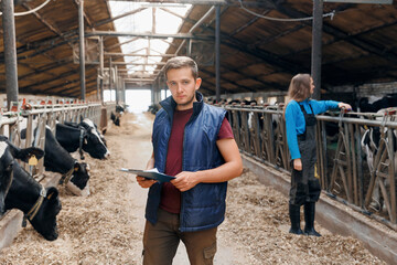 Portrait young man farmer with clipboard checking cows in cowshed on dairy farm, background vet doctor control health of cattle. Concept modern agriculture business industry, farming animal husbandry