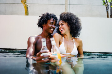 A young couple of African descent enjoying a sunny day at a luxurious poolside, sitting at the...
