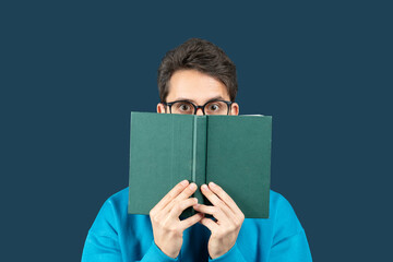 A young man sits against a solid dark blue background, holding an open book in front of his face....