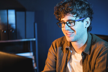 Smiling young businessman focusing on pc for creative startup project job at neon light modern office. Thinking marketing network online planning of creator to market channel design concept. Gusher.