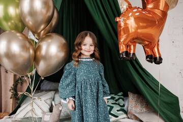 Child birthday party celebration. Happy little girl 4-5 years in stylish rustic style dress stands on bed decorated with baldachin and colorful foil balloons, smiles and looks at camera at bedroom © Andriy Medvediuk