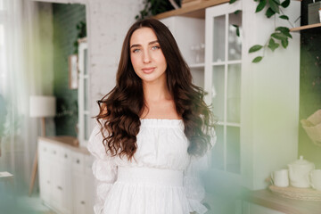 Women's day. Romantic attractive young woman 20-25 years with long curly hair in white dress is poses with crossed hands and looks at camera at modern stylish interior of home kitchen on background.