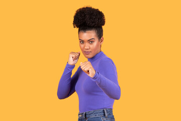 A young woman stands confidently with her fists raised, showcasing a fighting stance. She wears a purple sweater and denim jeans, highlighting her focus and determination