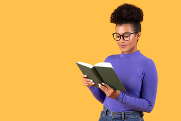 A young woman with curly hair styled in a bun is deeply focused on reading a book. She wears...