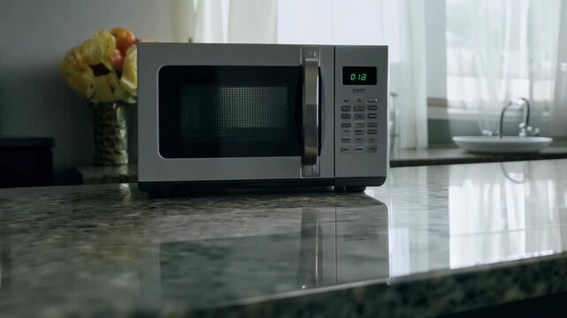 A silver microwave sits on a countertop in a kitchen. The microwave is turned off and has a digital display. The kitchen is well-lit and has a clean, modern look