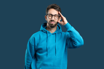 A young man is expressing discomfort while wearing a blue hoodie. He has glasses on and is standing against a solid background. His hand is placed on his forehead