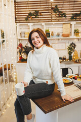 A cute woman in a white knitted sweater is sitting with coffee in a cozy kitchen with Christmas decorations. Kitchen in New Year's style.