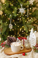 Christmas background. Cup of hot chocolate with marshmallows, christmas ornaments and gift, cozy knitted sweater and fir branches against stylish tree with lights. Merry Christmas. Winter hygge