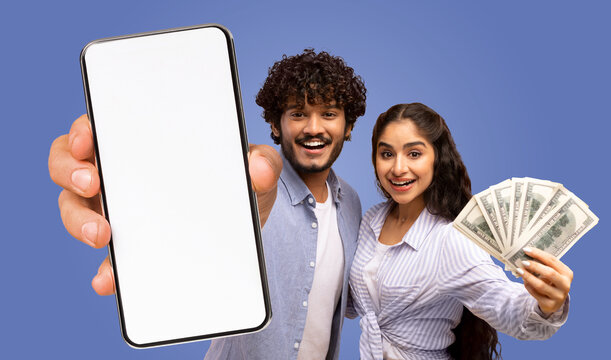 A young Indian couple is holding a smartphone with a blank screen and a fan of money, both facing the camera and smiling.