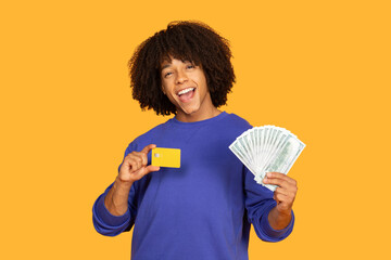 A cheerful young man is showcasing a handful of cash in one hand and a credit card in the other. The vibrant yellow background accentuates the excitement