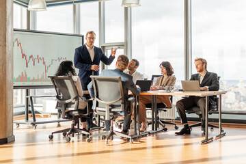 A group of business professionals are gathered around a conference table in a modern office. A presenter stands in front of the group, pointing to a large screen displaying a financial chart