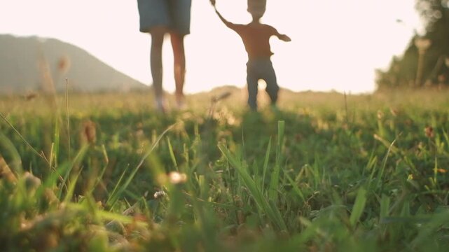 Caring young mother supports little son to learn walking forward, toddler growth and development concept. Selective focus of cute baby taking first steps in park on meadow grass at warm sunset