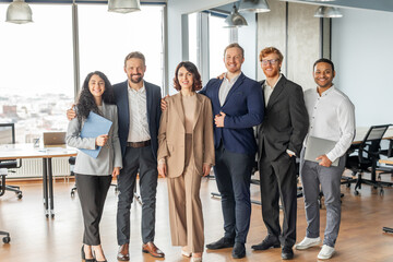 A group of six diverse business professionals stand together in a modern office setting. They are all smiling and appear to be in a good mood. The team is comprised of two women and four men