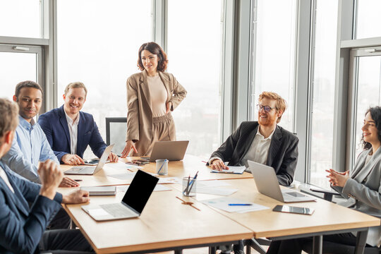A diverse team of business professionals are gathered around a conference table in a modern office, engaging in a lively discussion. The woman standing at the head of the table, leading the meeting