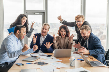 A team of five business professionals, dressed in suits and business casual attire, gather around a conference table in an office setting. They are all excitedly celebrating a success