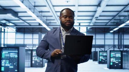 IT engineer working on laptop in modern data center, overseeing server operations, system...