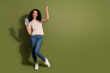 Full size photo of ecstatic girl dressed beige t-shirt hold smartphone raising fist win lottery isolated on khaki color background