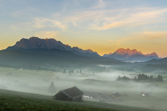 Mountain meadows with small huts in light ground fog and Zugspitz massif in the background at sunrise, Kr&uuml;n, Bavaria, Germany, Europe