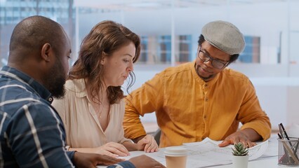 Professional clients and their real estate agent evaluating blueprints for the high class modern space, discussing leasing contract terms before signing. Startup business development. Camera B.