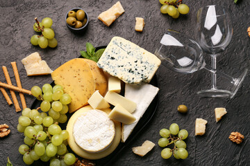 Tray with different types of tasty cheese, grapes and wine glasses on dark background