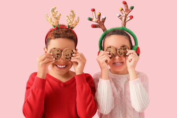 Funny little children in reindeer horns with cookies on pink background