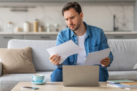A man sits on a white couch in a living room, reviewing documents. He has a concerned look on his face as he carefully looks over the papers while sitting in front of an open laptop